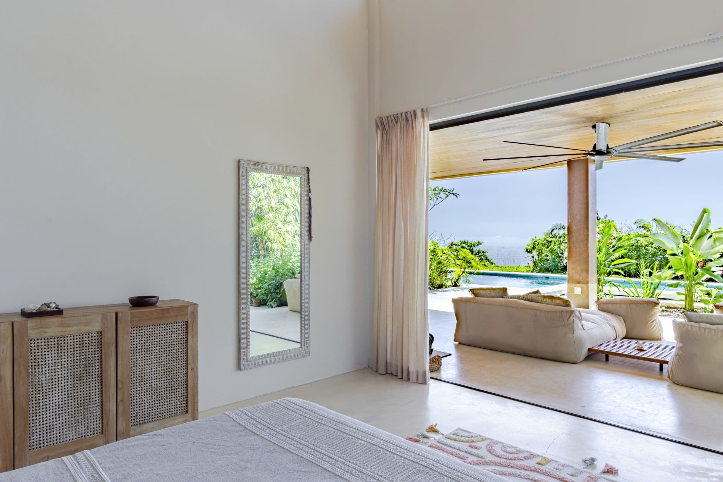 View from a bedroom through open glass walls to the lounge, pool and ocean horizon