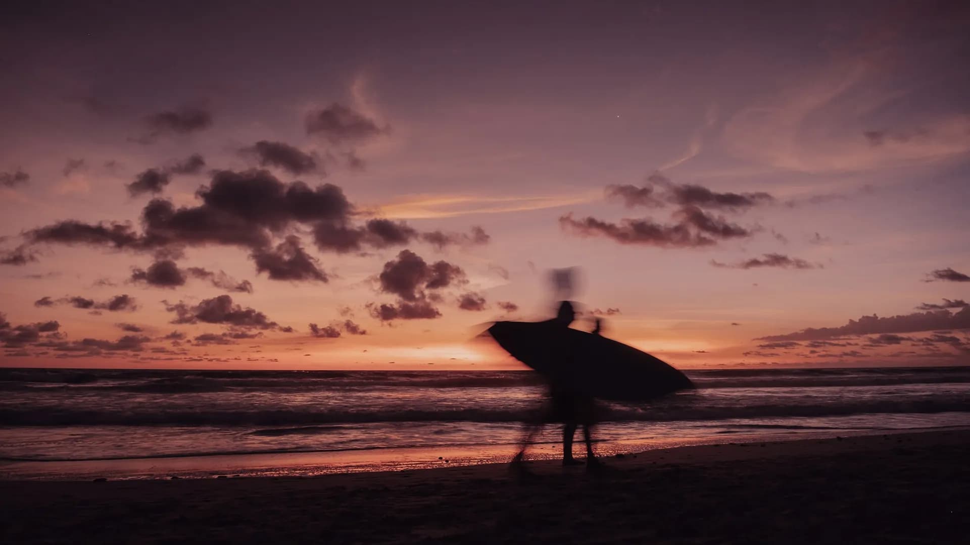 A surfer with a board silhouetted against a violet and orange sunset at Santa Teresa Beach.