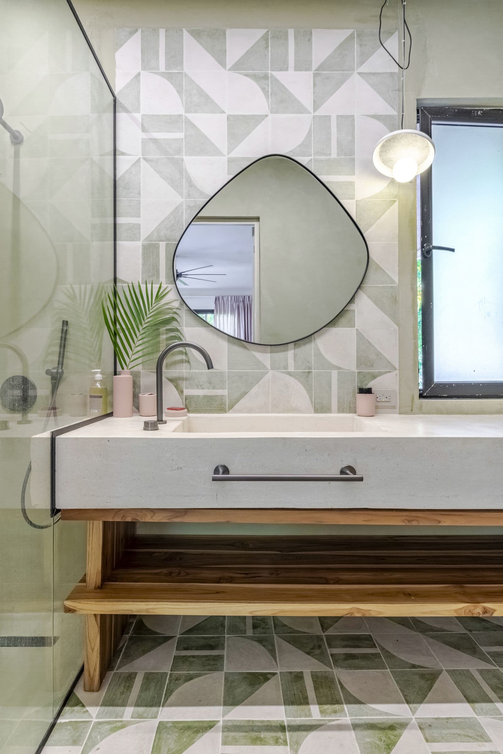 Bathroom with green geometric tiles, a pebble-shaped mirror and a basin counter on an open wooden shelf