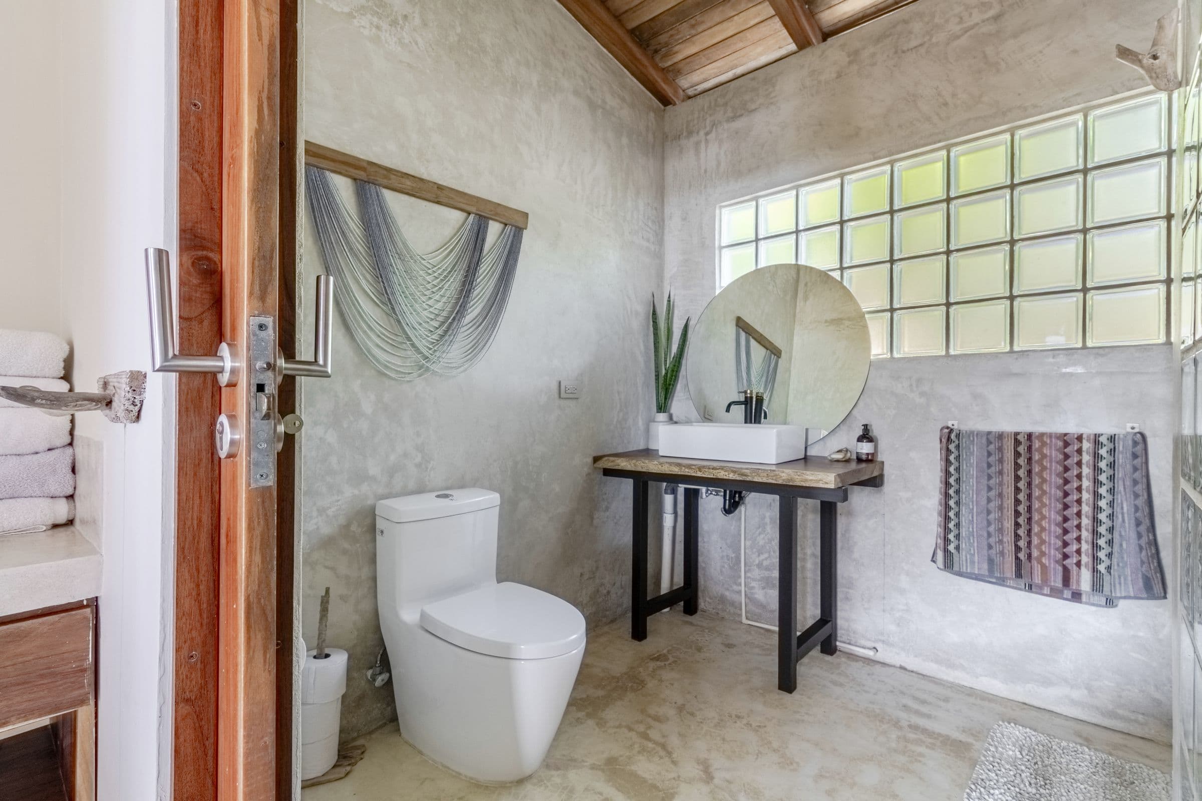 Bathroom with textured grey walls, a glass-block window, a round mirror and a vessel sink on a wooden-topped stand