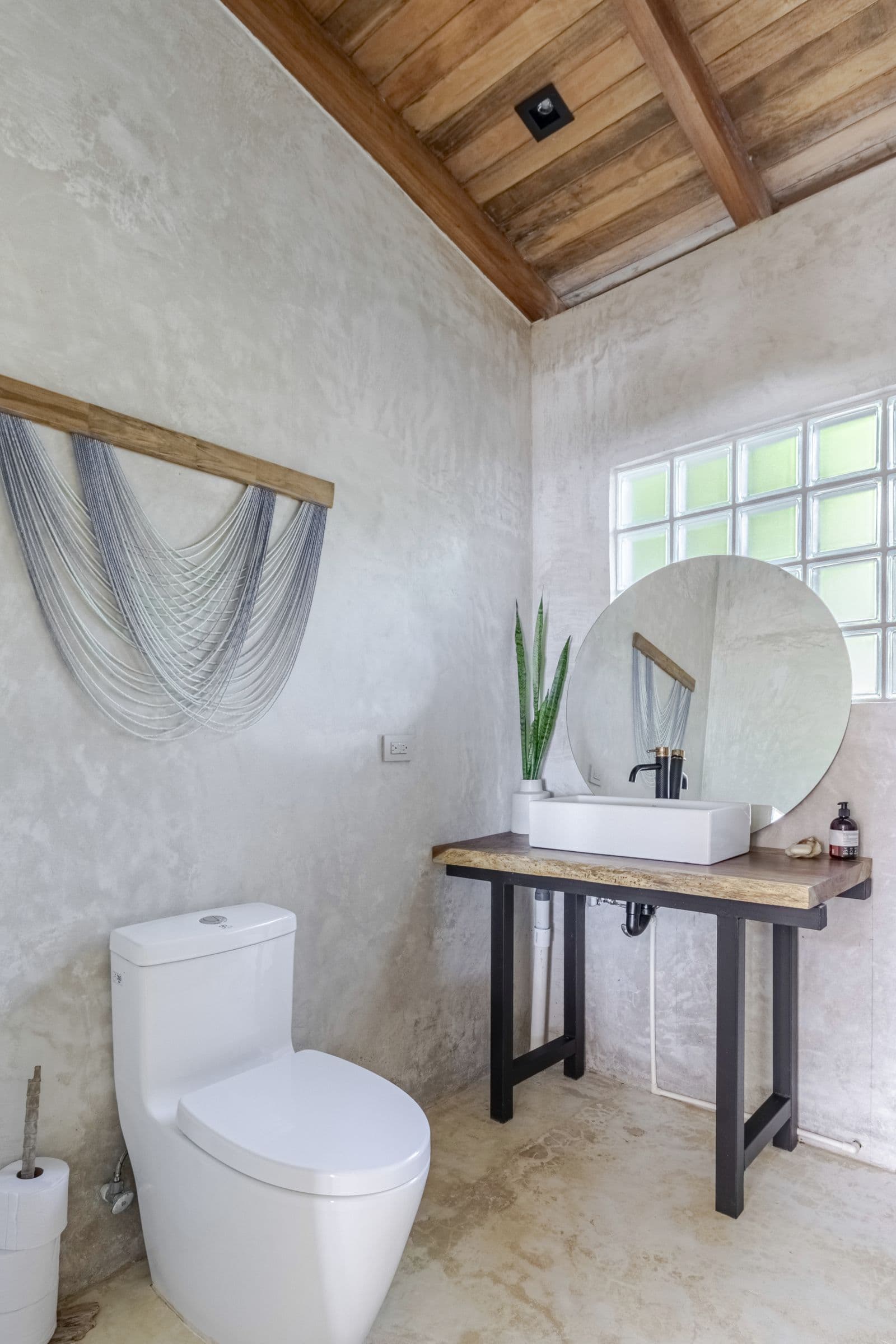 Bathroom with a woven wall hanging, a round mirror and a vessel sink beneath a sloping timber ceiling