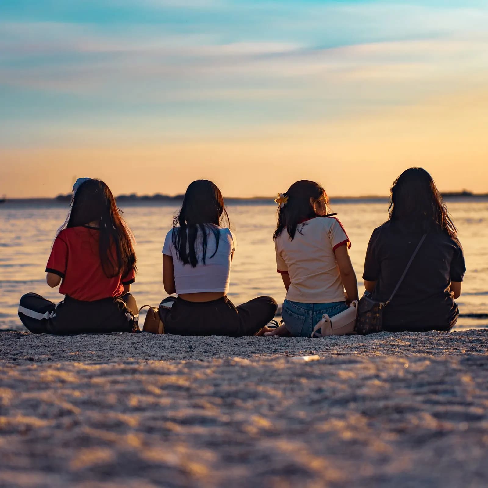 Four friends sit side by side on the sand watching the sunset over the water.