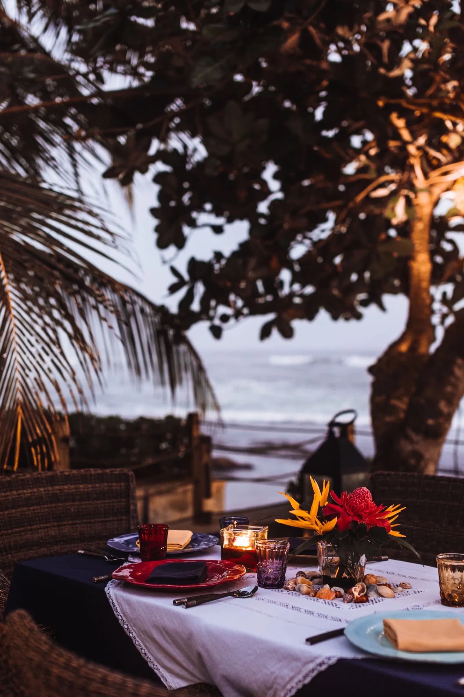 A candlelit table with tropical flowers and white linen overlooks the sea beneath coastal trees at dusk.