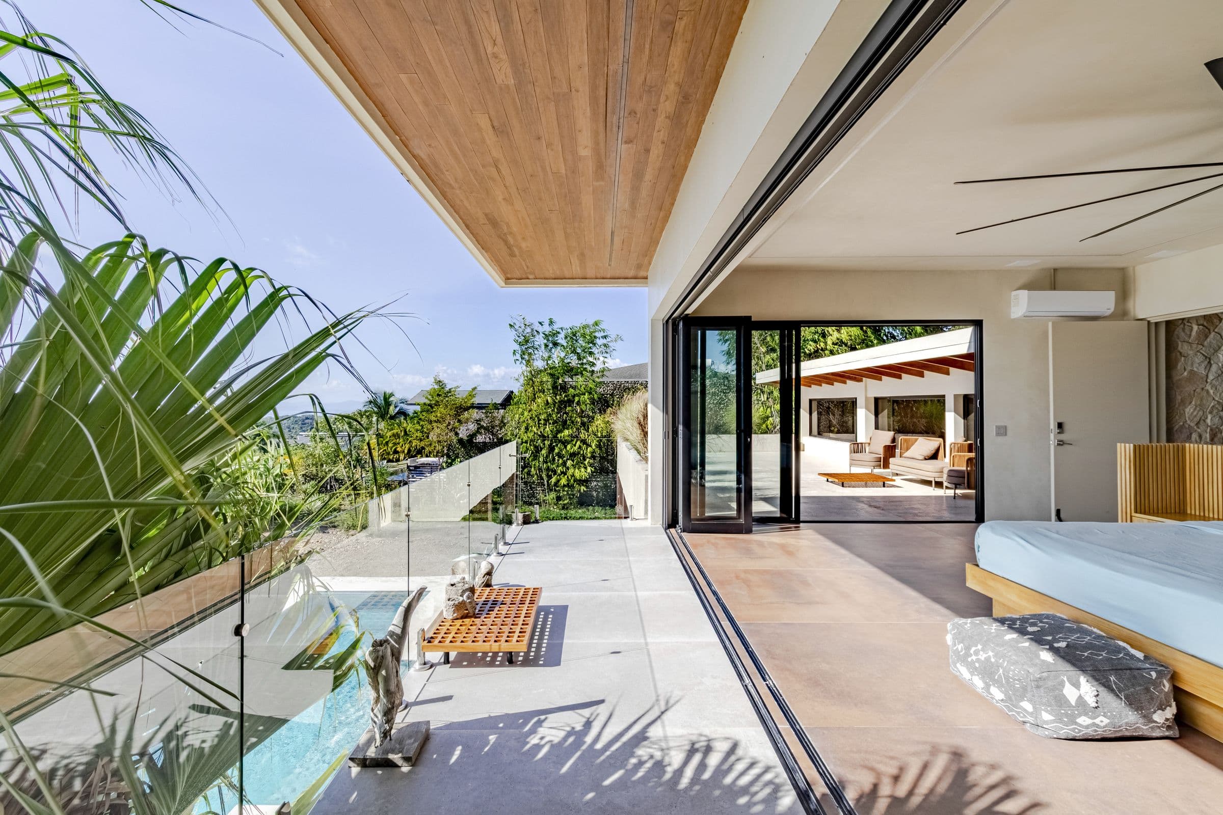 Upper-floor bedroom balcony with a glass railing overlooking the pool and tropical garden