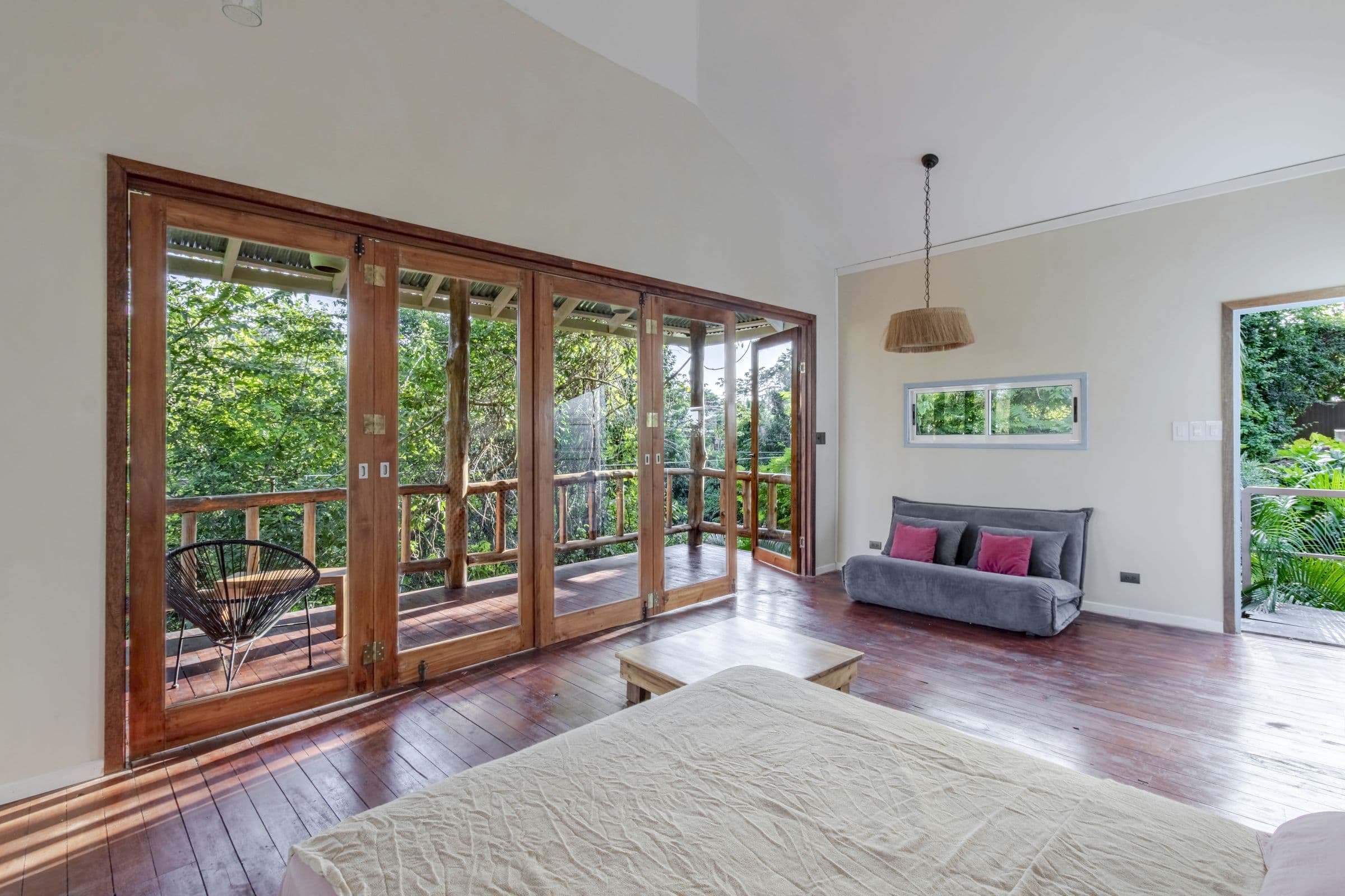 Inside the Surf Shack: a bed, a grey sofa bed and folding wooden doors open to a balcony in the trees